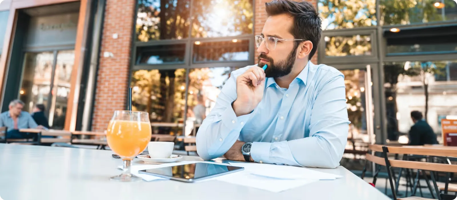 a man is sitting in a restaurant on the street with a thoughtful look and a pen in his hand, a tablet and papers in front of him