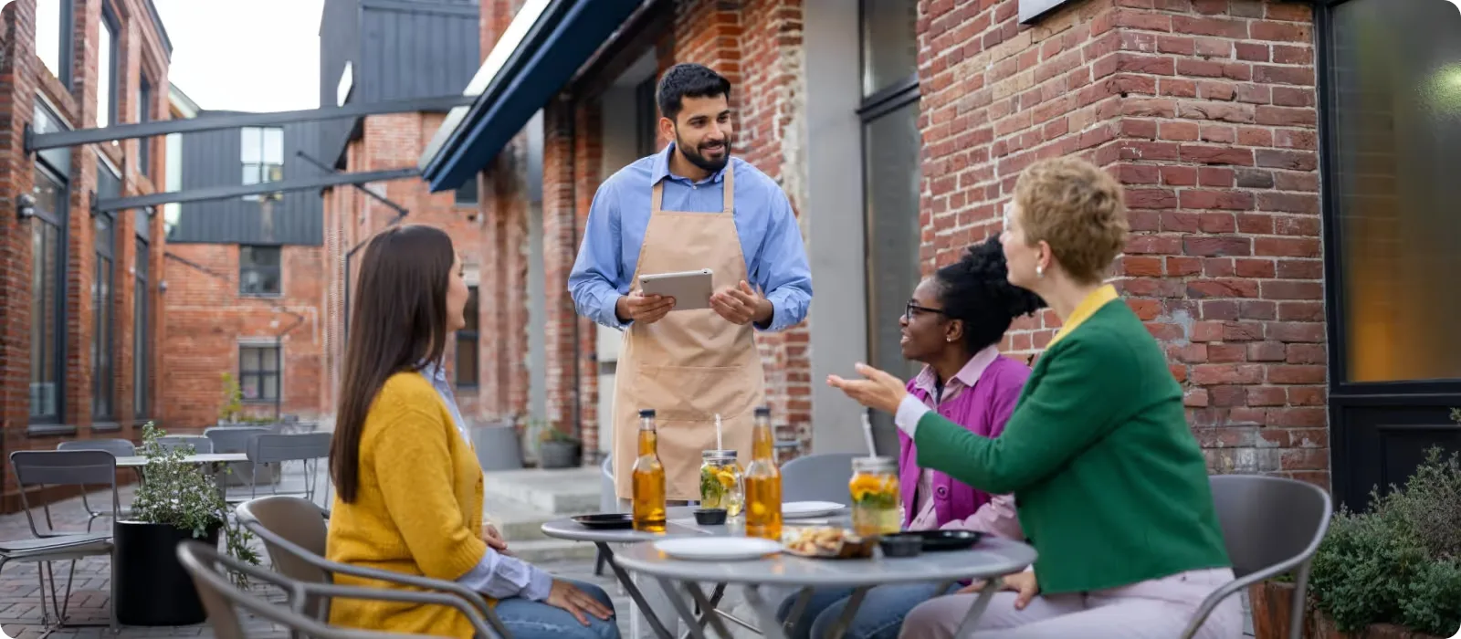 Three women are sitting outside in a restaurant, a waiter with a tablet tells them the menu
